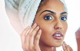 A woman with a towel wrapped around her hair is gently touching her face, showcasing a natural skincare routine. She has a glowing complexion and is looking directly at the camera, conveying a sense of confidence and self-care. The background is softly blurred, emphasizing her features and the importance of skin health. This image highlights beauty, wellness, and the significance of daily skincare practices.
